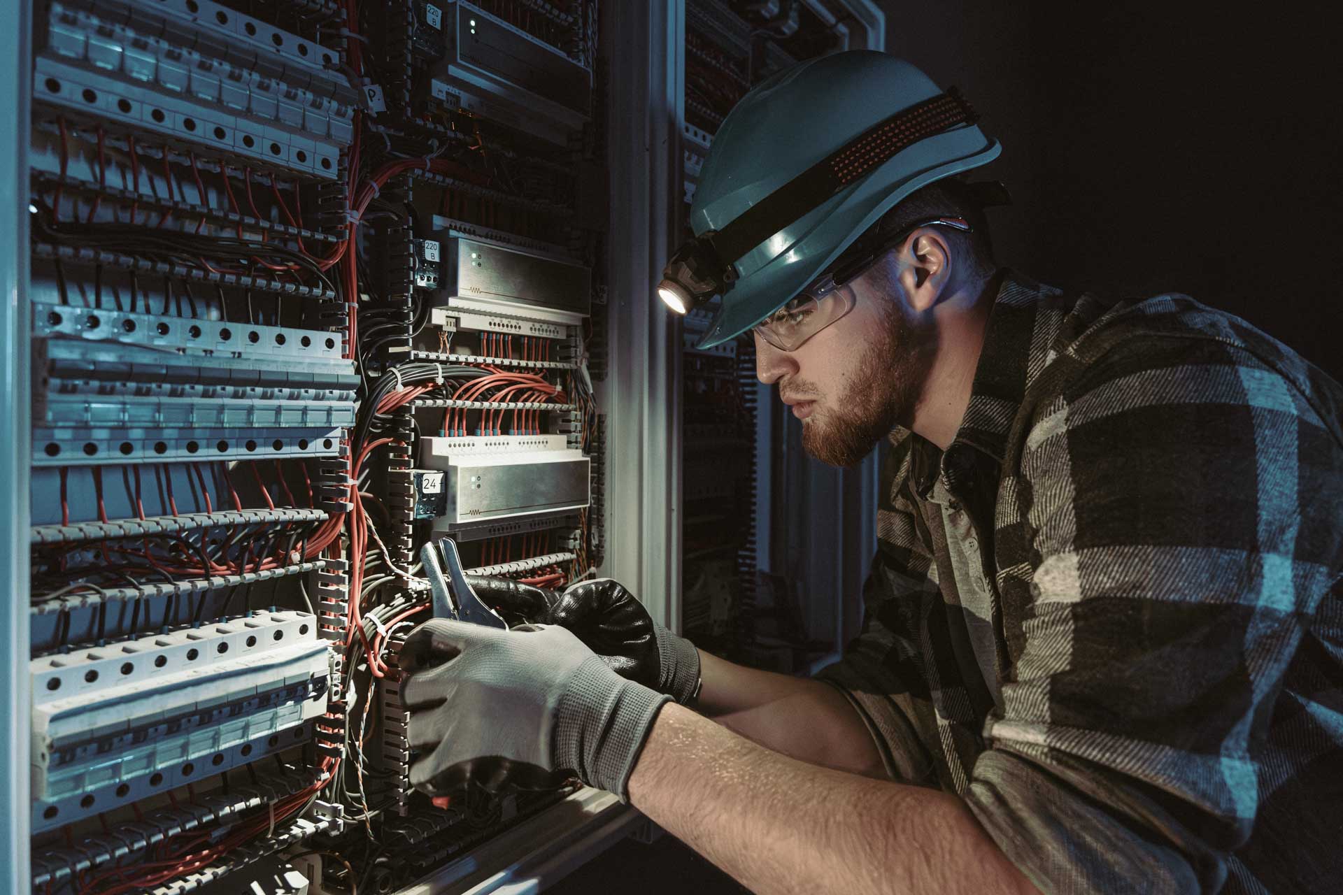 A Male Electrician Works In A Switchboard In Overalls Against The Backdrop Of Emergency Lighting.